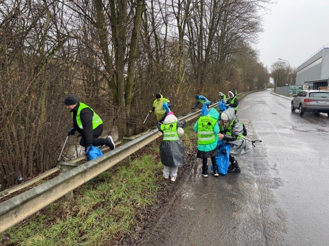 Man sieht Menschen und Kinder die entlang der Selz Müll bei Regenwetter sammeln. Die Personen tragen Warnwesten, Regenschutz und Müllbeutel während sie den Müll sammeln.
