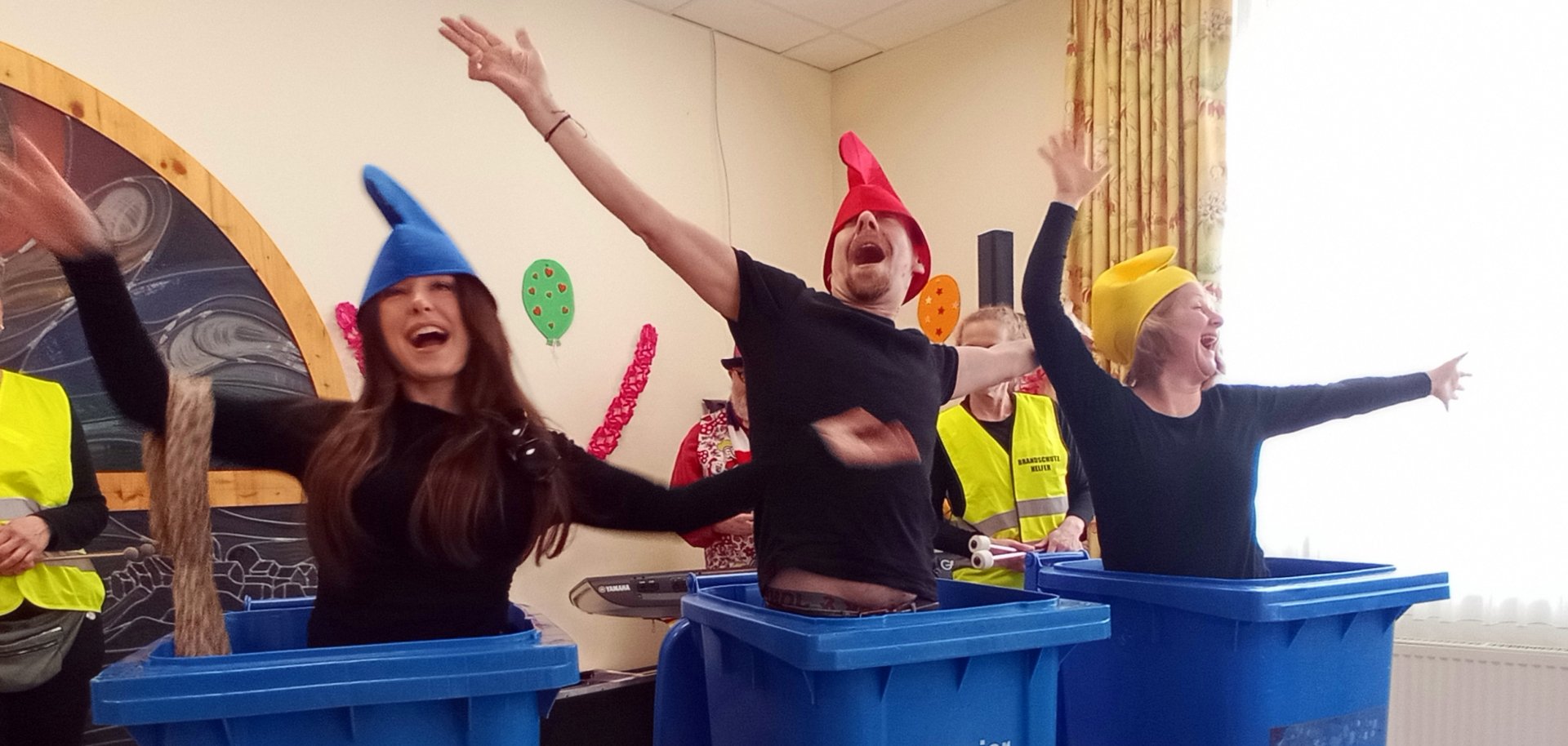 Man sieht 3 Menschen mit schwarzen Shirts und bunten Mützen, die in 3 blauen Altpapiermülltonnen stehen und dabei tanzen.