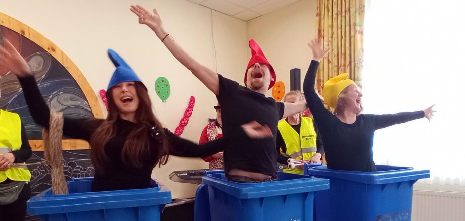 Man sieht 3 Menschen mit schwarzen Shirts und bunten Mützen, die in 3 blauen Altpapiermülltonnen stehen und dabei tanzen.