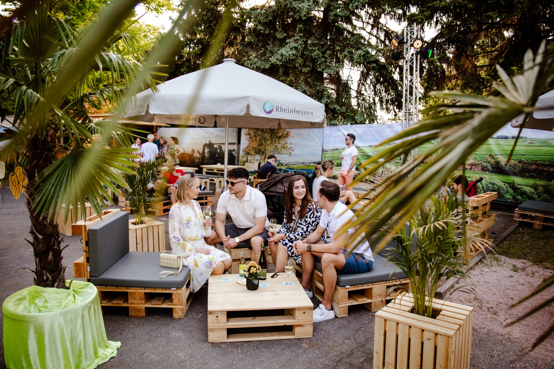 Auf dem Bild sieht man Menschen die sitzend das Weinfest "Scheutime" genießen und dabei Wein trinken und sich unterhalten, desweiteren sind ein Sonnenschirm, einige Palmen und Pflanzen zu sehen.