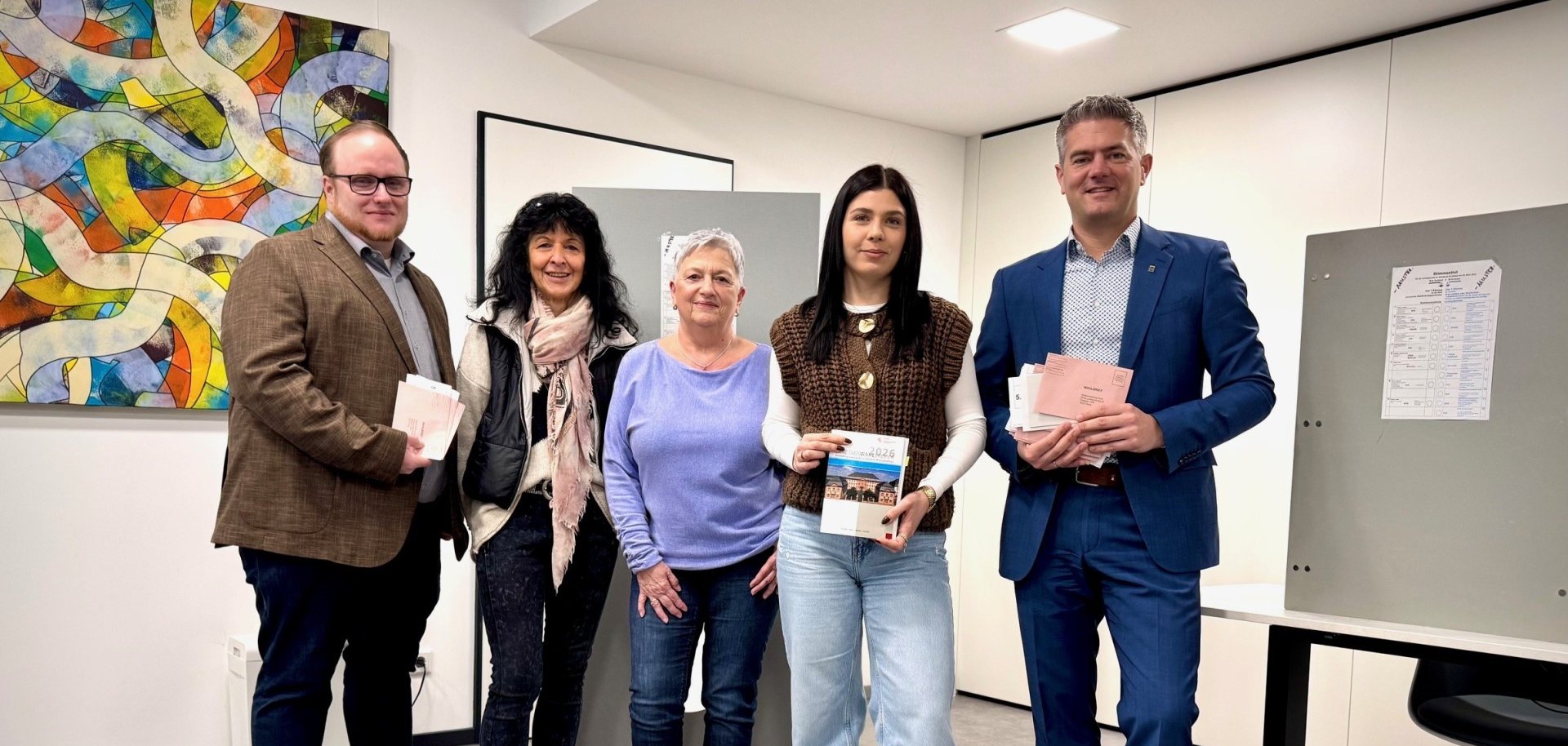 Man sieht Bürgermeister Steffen Jung und Mitarbeiteter der Stadt Alzey im Briefwahlbüro stehen die, die Briefe für die Landtagswahlen 2026 halten und in die Kamera lächeln, vor ihnen Steht eine Urne auf einem Tisch.