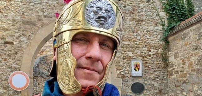 Man sieht einen als Ritter verkleideten Mann vor einer Mauer der einen Werbeflyer des Römerfestes hält.