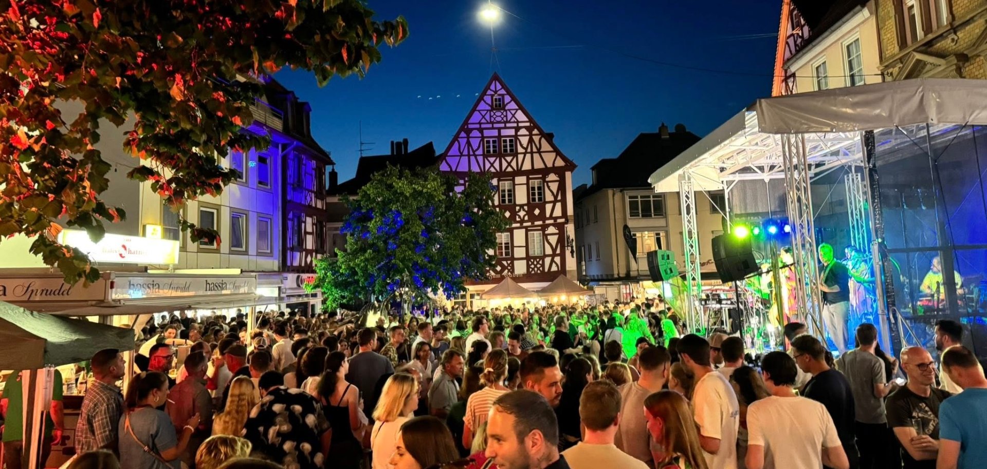 eine Menschenmenge steht bei Nacht vor mehreren Ausschankständen und vor einer Bühne