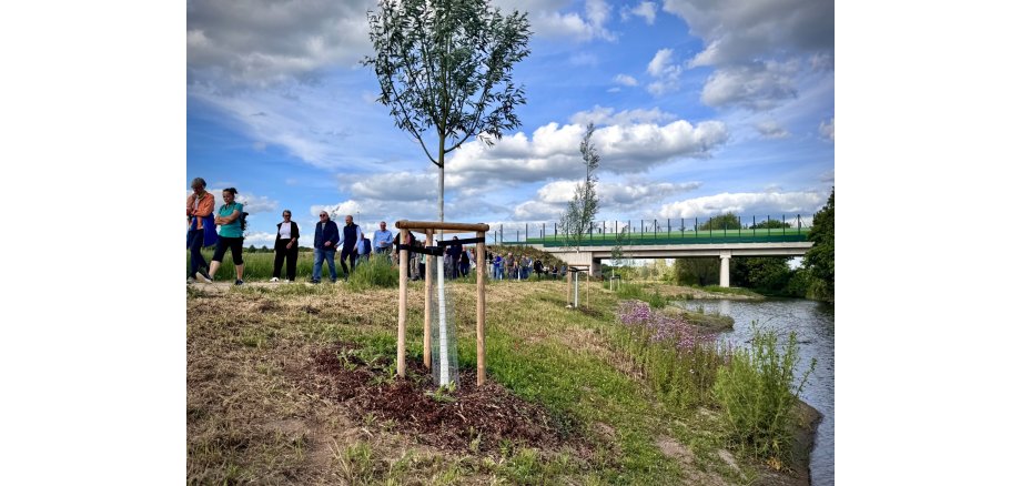 rechts im Bild ist ein Fluss abgebildet, links daneben läuft eine Gruppe von Menschen auf einem Feldweg