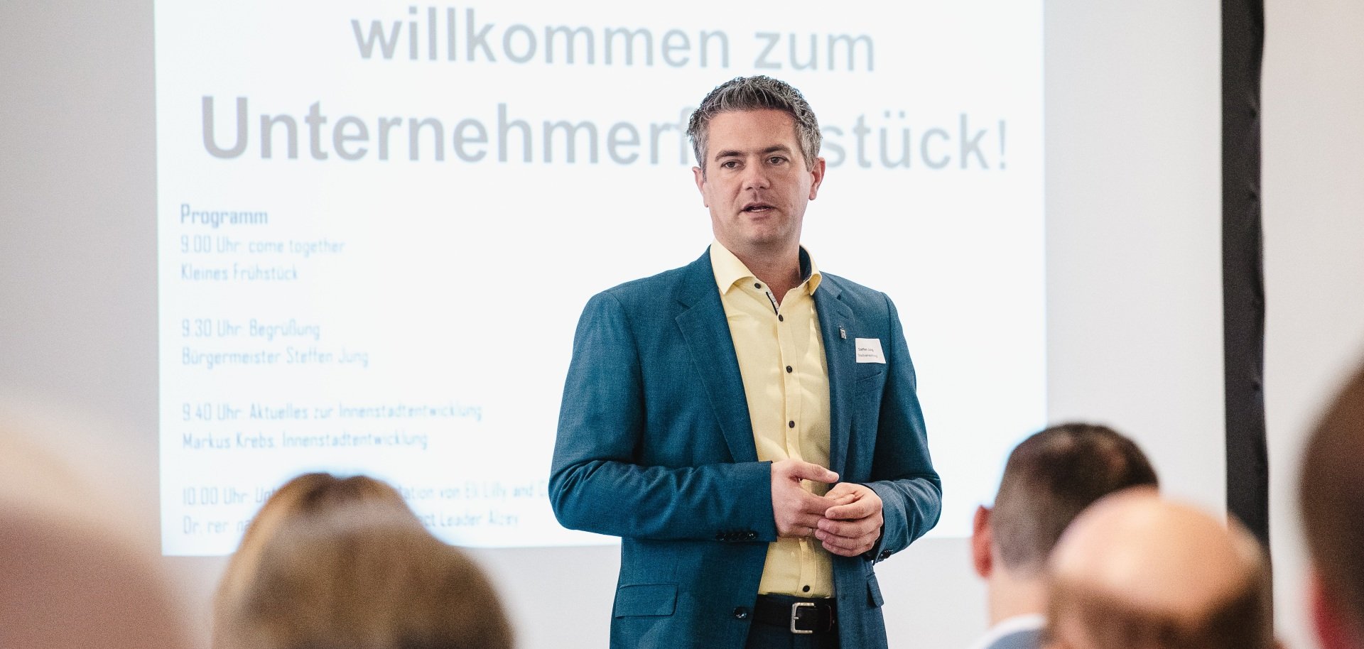 Man sieht im Vordergrund Bürgermeister Steffen Jung der vor der Präsentation: "Herzlich wilkommmen zum Unternehmerfrühstück" steht und die Teilnehmer begrüßt.