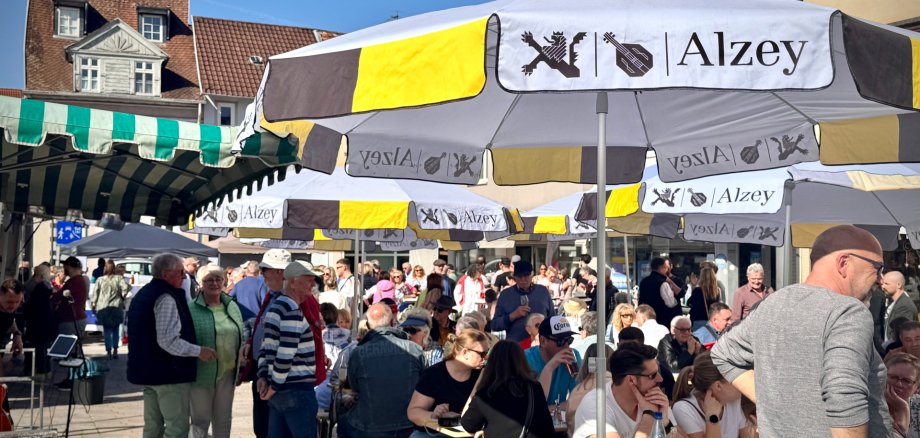 Man sieht den Wochenmarkt, Menschen die sitzen, stehen, trinken und sich unterhalten.