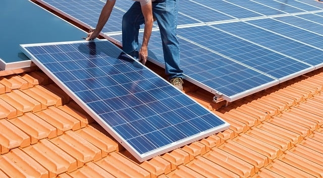 auf dem Foto ist ein Mann auf einem roten Ziegeldach abgebildet, der grade Solarpaneele darauf montiert