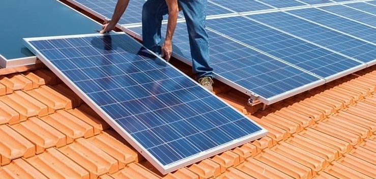 auf dem Foto ist ein Mann auf einem roten Ziegeldach abgebildet, der grade Solarpaneele darauf montiert