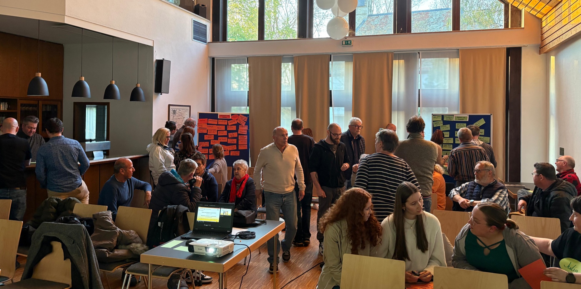 Einige Menschen laufen durch den Raum, andere sitzen gemeinsam an Tischen. In den Ecken im Hintergrund sind zwei Pin-Boards aufgestellt, an denen einige Zettel mit Aufschriften hängen.