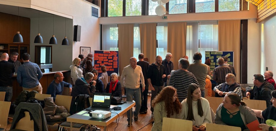 Einige Menschen laufen durch den Raum, andere sitzen gemeinsam an Tischen. In den Ecken im Hintergrund sind zwei Pin-Boards aufgestellt, an denen einige Zettel mit Aufschriften hängen.
