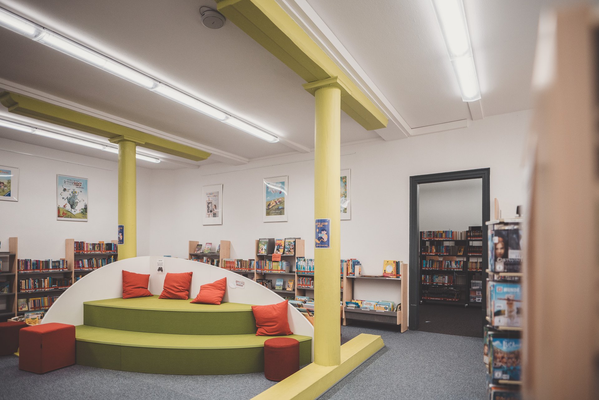eine Bücherei von innen mit einem Sitzbereich in den Farben orange und grün, im Hintergrund sind Bücherregale, gefüllt mit vielen Büchern zu sehen