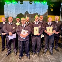 Einige Feuerwehrmänner stehen mit Bürgermeister Jung (links) auf einer Bühnme und halten Urkunden in der Hand und schauen in Richtung Kamera.