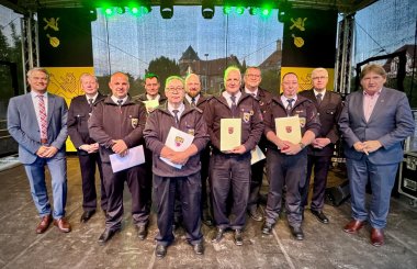 Einige Feuerwehrmänner stehen mit Bürgermeister Jung (links) auf einer Bühnme und halten Urkunden in der Hand und schauen in Richtung Kamera.