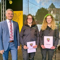 Auf einer Bühne stehen zwei Frauen in Uniform und halten jeweils eine Urkunde in der Hand. Bürgermeister Steffen Jung steht links daneben. Alle lachen in die Kamera.