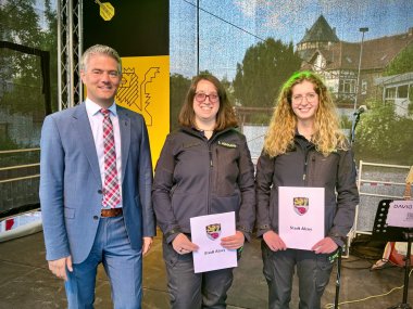 Auf einer Bühne stehen zwei Frauen in Uniform und halten jeweils eine Urkunde in der Hand. Bürgermeister Steffen Jung steht links daneben. Alle lachen in die Kamera.