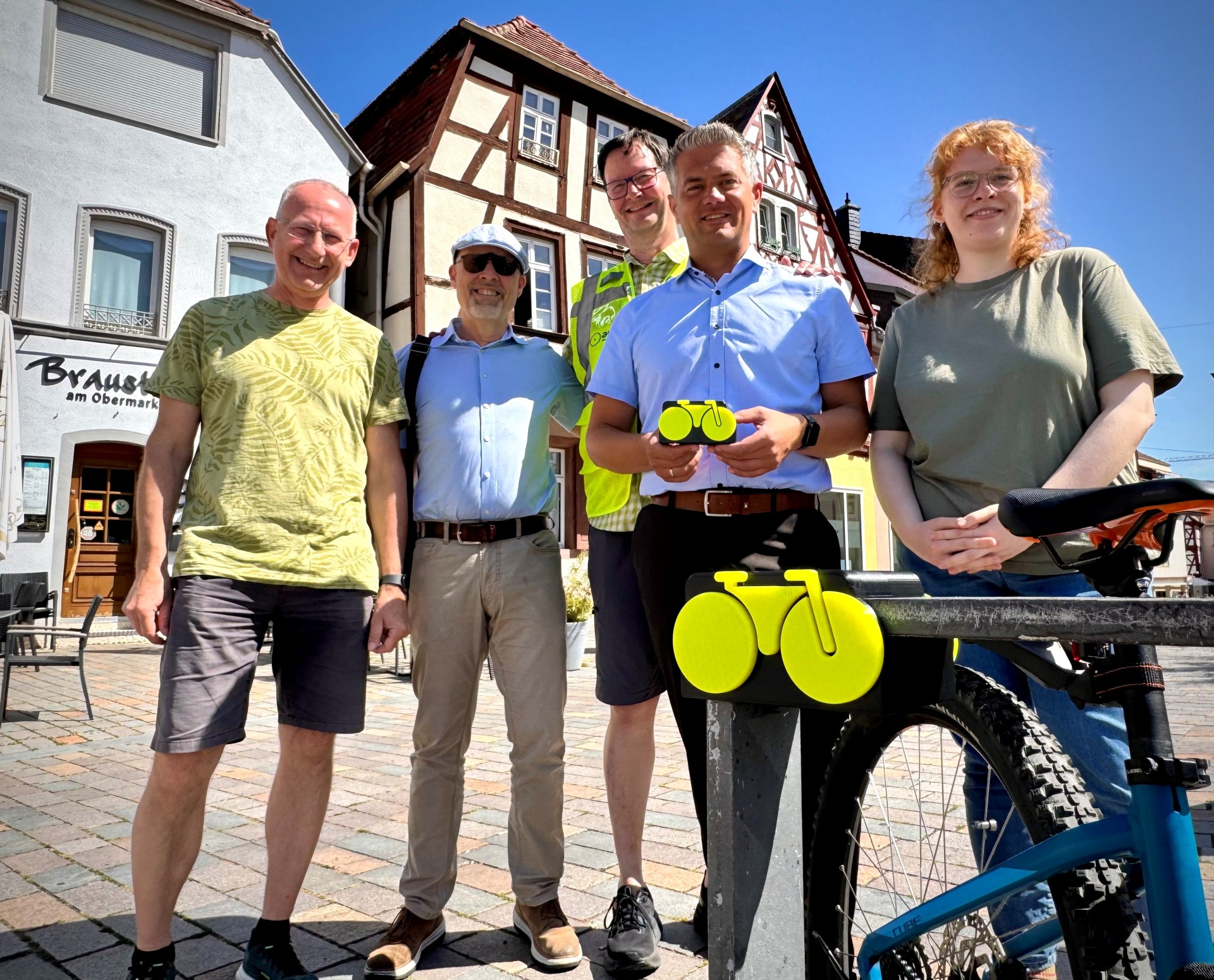 fünf Personen stehen vor der Kamera und lachen in diese, mittig steht Bürgermeister Steffen Jung und hält ein kleines gelbes Modellfahrrad in die Kamera