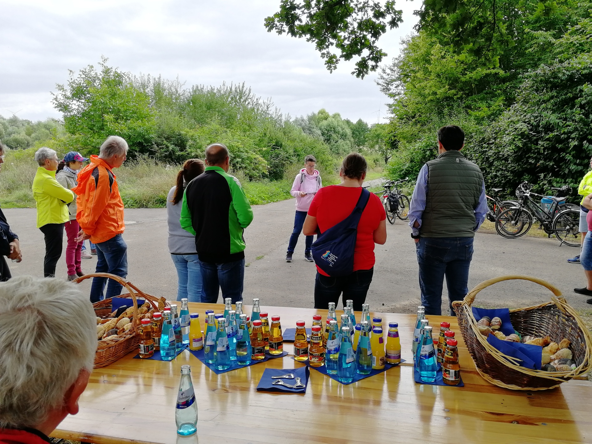 Im Vordergrund steht ein Tisch, auf diesem etwas Laugengebäck in Brotkörben und Wasserflaschen, Apfelsaft und Orangensaft. Dahinter steht eine Gruppe von Menschen in Fahrradkleidung, die alle einer Frau zuhören. Hinter dieser Frau stehen einige Fahrräder abgestellt.