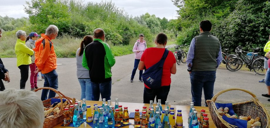 Im Vordergrund steht ein Tisch, auf diesem etwas Laugengebäck in Brotkörben und Wasserflaschen, Apfelsaft und Orangensaft. Dahinter steht eine Gruppe von Menschen in Fahrradkleidung, die alle einer Frau zuhören. Hinter dieser Frau stehen einige Fahrräder abgestellt.