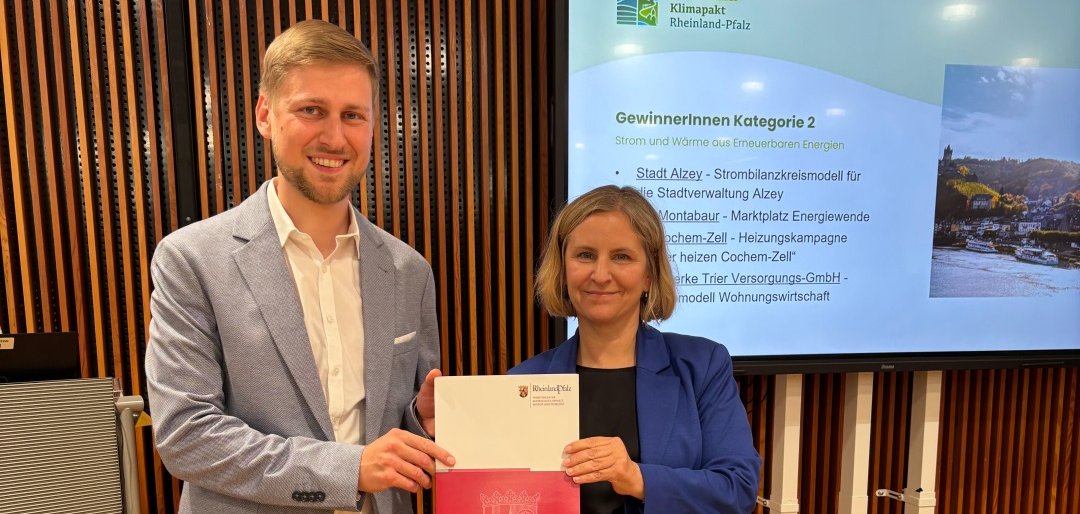 Vor der Kamera stehen zwei Personen, eine Frau und ein Mann, die gemeinsam eine Urkunde in der Hand halten und in die Kamera zeigen. Beide sind schick angezogen. Im Hintergrund ist ein Bildschirm mit der Aufschrift Gewinner Klima Kategorie 2