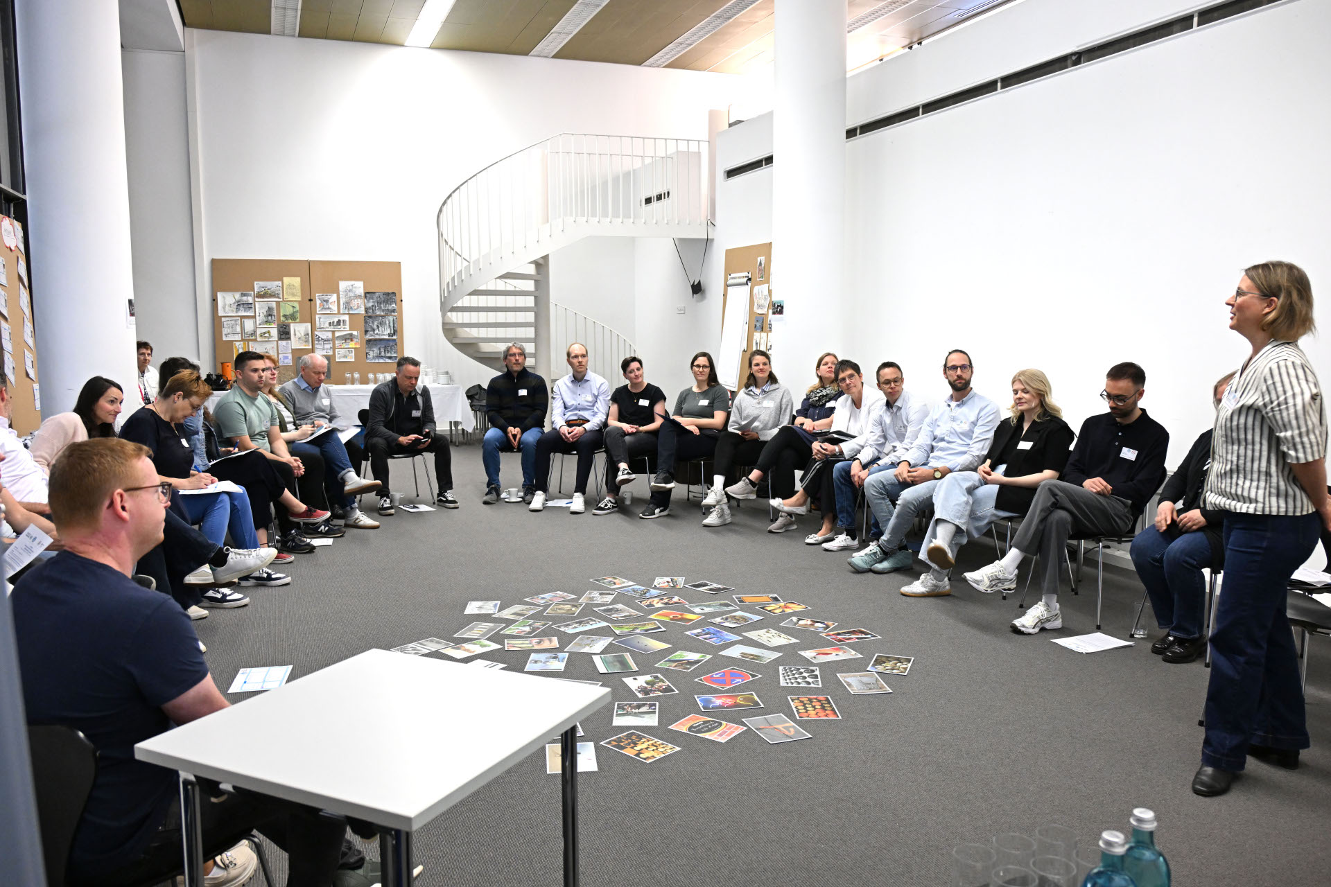 In einem Besprechungsraum sitzen mehrere Personen in einem Stuhlkreis. In ihrer Mitte befinden sich bunte Karten auf dem Boden, die alle eine Aufschrift haben.