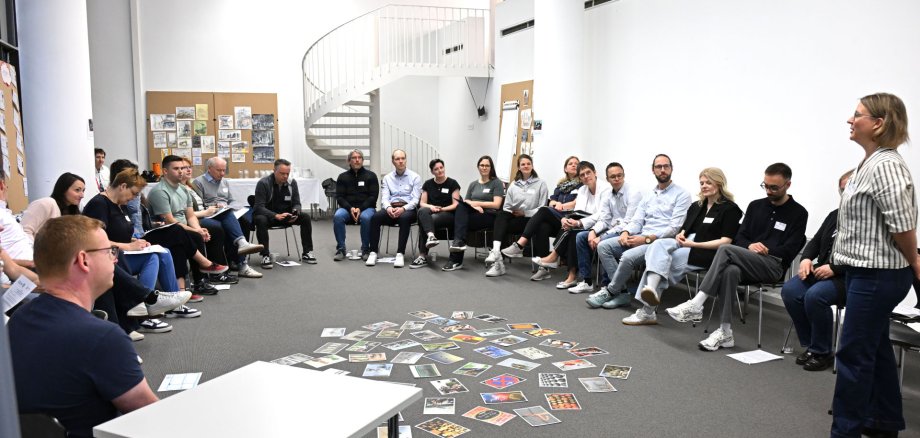In einem Besprechungsraum sitzen mehrere Personen in einem Stuhlkreis. In ihrer Mitte befinden sich bunte Karten auf dem Boden, die alle eine Aufschrift haben.