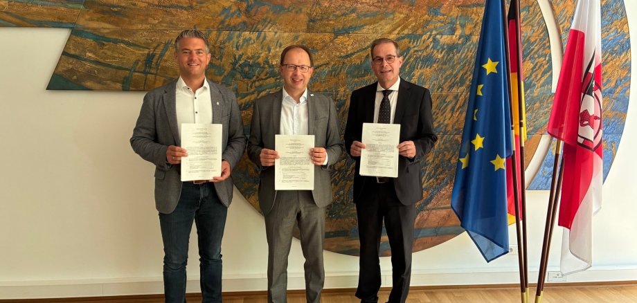 drei Männer in Anzug stehen vor einer Wand und halten alle jeweils einen Vertrag in der Hand und lachen in die Kamera