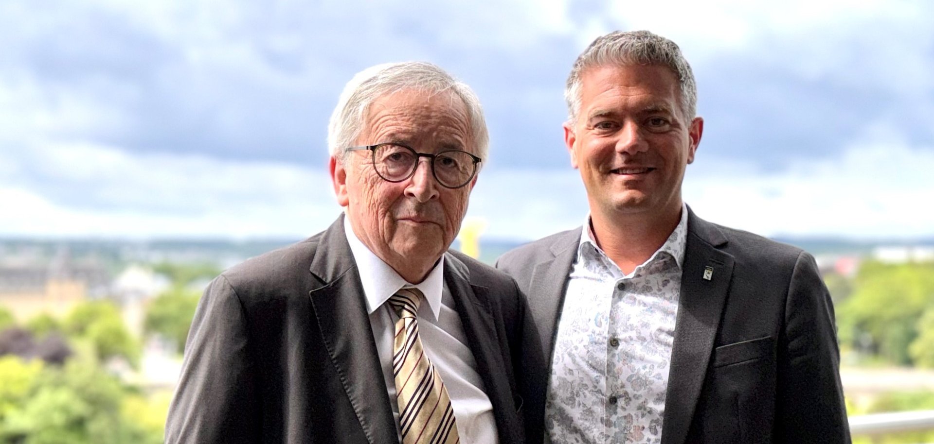 zwei Männer in Anzug stehen nebeneinander vor einer Grünfläche im Hintergrund und lachen in die Kamera. Der rechtsstehende Mann ist Bürgermeister Steffen Jung, links daneben Jean-Claude Juncker
