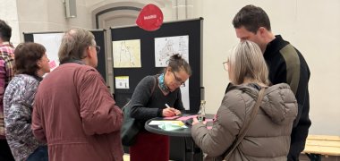 Innenstadt Nord - Auftaktveranstaltung, Stand zum Thema "Mobilität", zu sehen sind Menschen die an einem Tisch stehen und  eine Frau die etwas schreibt.