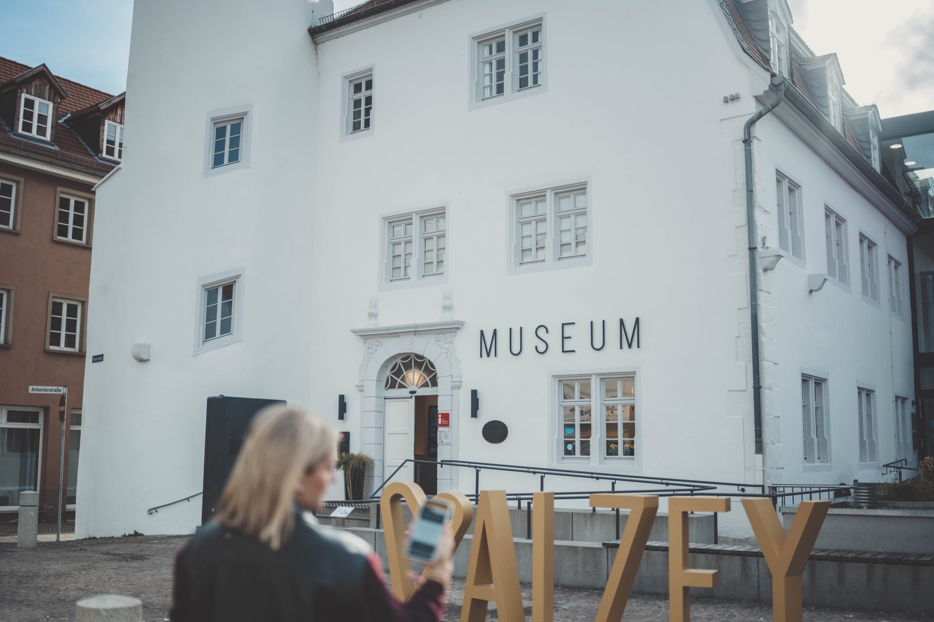 Man sieht das Museum im Hintergrund, den Fotopoint Alzey, und ein Handy mit der geöffneten Stadt-App. (Alzey-App)
