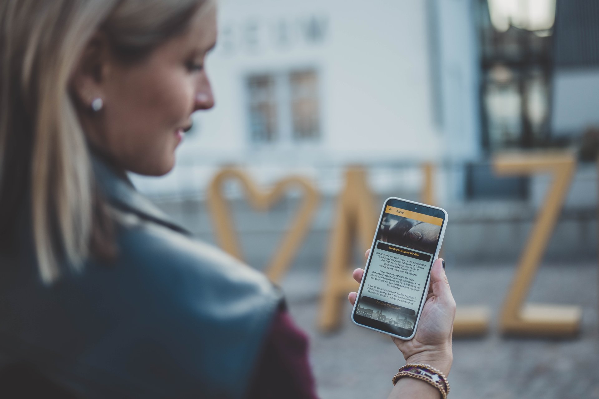Man sieht das Museum im Hintergrund, den Fotopoint Alzey, und ein Handy mit der geöffneten Stadt-App. (Alzey-App)