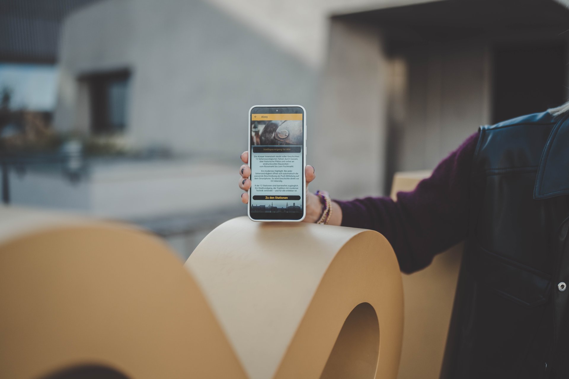 Man sieht das Museum im Hintergrund, den Fotopoint Alzey, und ein Handy mit der geöffneten Stadt-App. (Alzey-App)