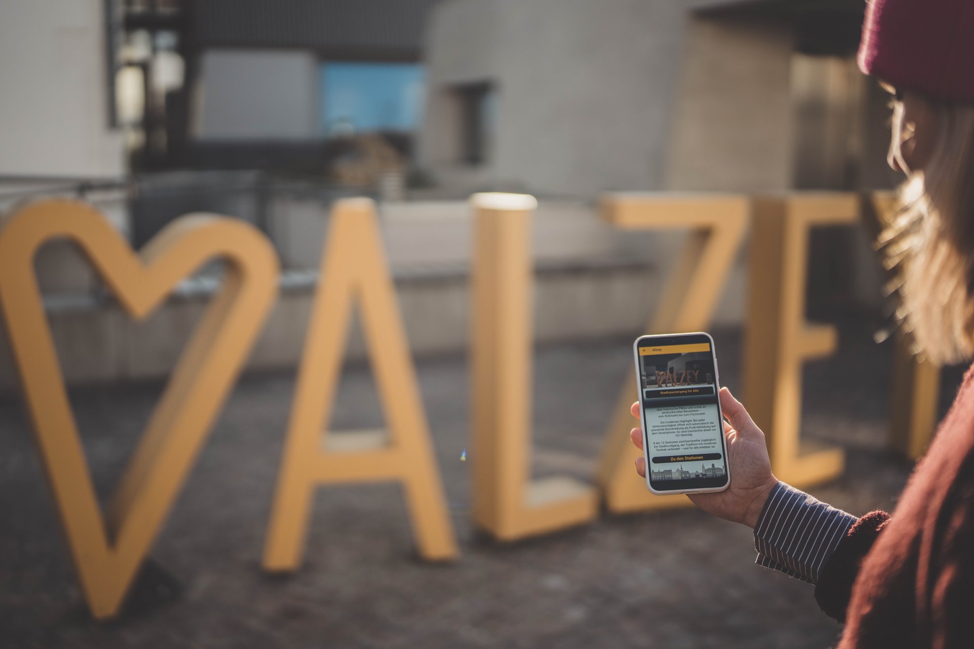 Man sieht das Museum im Hintergrund, den Fotopoint Alzey, und ein Handy mit der geöffneten Stadt-App. (Alzey-App)